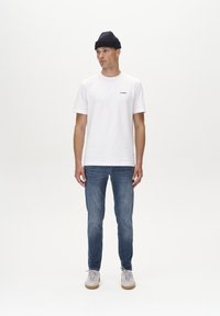 Weißes Baumwoll-T-Shirt mit einem kleinen schwarzen Logo, kombiniert mit blauen Jeans und hellgrauen Sneakers. Das Modell trägt eine dunkle Strickmütze.