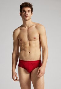 Culottes en coton rouge avec un ajustement confortable, dotées d'une large ceinture et de coutures décoratives. Le modèle se tient devant un fond gris neutre.