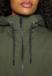 Grön huppjacka med framdragkedja, utrustad med svart och vita dragsko och en logga på bröstet. Slät textur, avslappnad design.