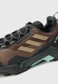 Braune und schwarze Outdoor-Schuh mit Mesh- und synthetischen Materialien, strukturierter Oberfläche, kontrastierenden drei Streifen und Gummisohle mit Profil.