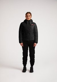 Giacca a vento nera con cappuccio, texture trapuntata, zip frontale e due tasche laterali con zip. Indossata con pantaloni e scarpe nere.