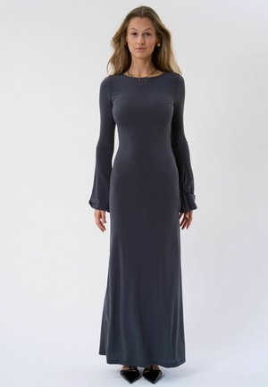 Frau mit langen Haaren, die ein eng anliegendes, langärmliges, bodenlanges dunkelgraues Kleid und schwarze spitze Schuhe trägt, vor einem schlichten weißen Hintergrund steht.