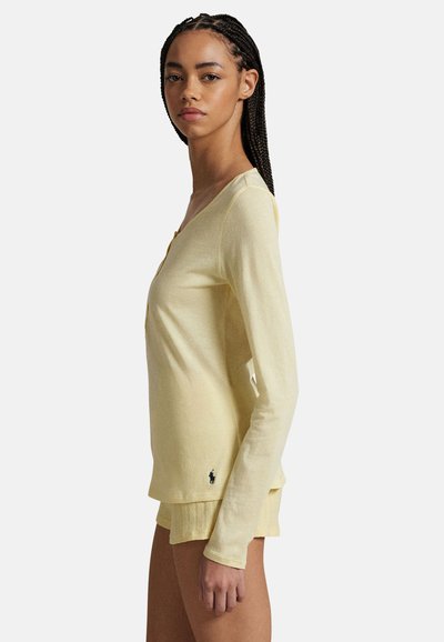 Jeune femme aux cheveux tressés portant un haut léger à manches longues jaune pâle et un short assorti, debout de profil devant un fond uni.