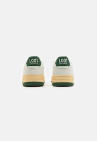 Sneakers bianchi in pelle con linguette verdi sul tallone e testo "L001 LACOSTE". Suola intermedia gialla e suola in gomma verde. Forma arrotondata, design minimale.