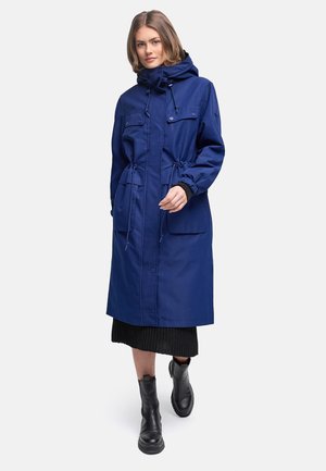 Ženska v dolg plašč z navy kapuco z žepi spredaj, črni midi krili in črnih gležnjarjih stoji na belem ozadju.