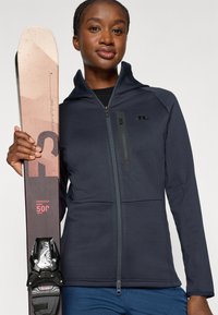 Chaqueta de cremallera azul marino con un cuello alto y un bolsillo en el pecho, sosteniendo esquís marrones y blancos con fijaciones negras con acentos blancos.
