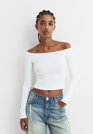 Top alb cu umeri goi și mâneci lungi, din material texturat în dungi, combinat cu blugi de culoare albastră, de talie înaltă, ușor decolorati, evidențiind un croi lejer și un stil casual.
