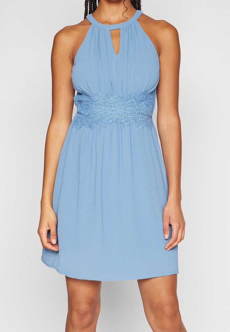 Mouwloze lichtblauwe jurk met halternek, open sleutelgat aan de voorkant en brede kanten tailleband, gedragen door een staand persoon.