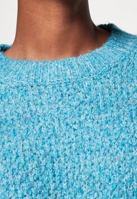 Stickad tröja i gradientnyanser av ljusblått med en texturerad, grov struktur. Har en ribbad rund halsringning.