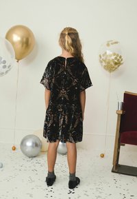 Melnas samta kleitas ar krāsainu mirdzošu ziedu rakstu, īsām piedurknēm un savilktu svārku. Stilizēta ar melnajām kurpēm un sudraba zeķēm.