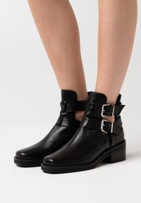 Bottes noires en cuir jusqu'aux chevilles avec des boucles en métal argenté, bout légèrement pointu et petit talon carré, présentant des découpes et une texture lisse.