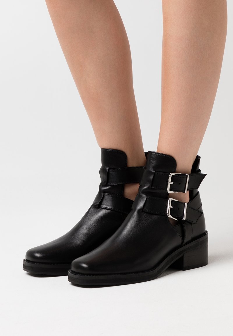 Bottes noires en cuir jusqu'aux chevilles avec des boucles en métal argenté, bout légèrement pointu et petit talon carré, présentant des découpes et une texture lisse.