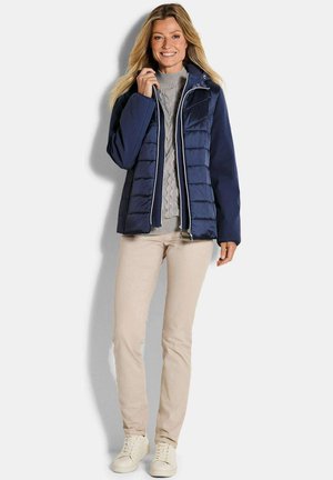 Frau lächelt, trägt eine marineblaue gesteppte Jacke über einem grauen Zopfmuster-Pullover, beige Hosen und weiße Turnschuhe, steht vor weißem Hintergrund.