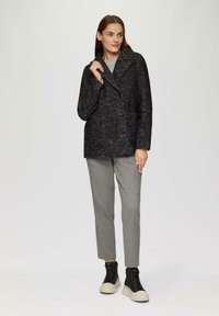 Abrigo negro tipo peacoat con un tejido texturizado, botones de doble botonadura y grandes solapas, combinado con pantalones grises y zapatillas altas.