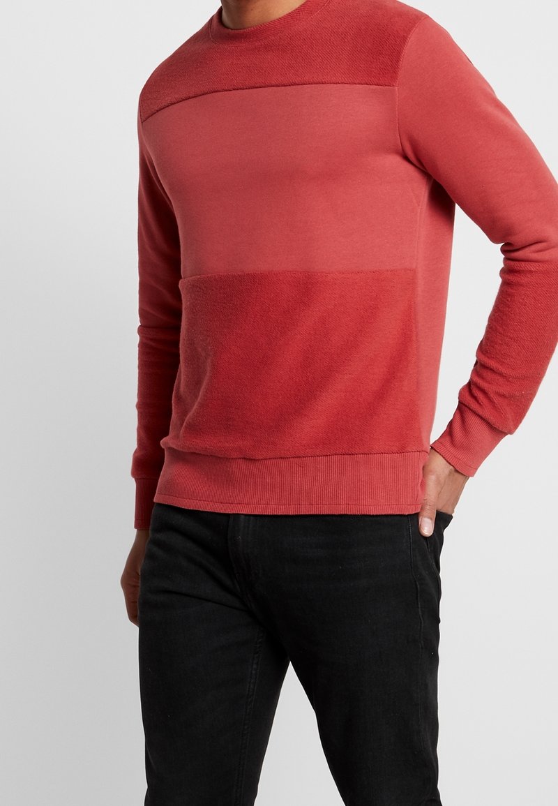 Homme portant un pull rouge bicolore à manches longues et un pantalon noir, une main dans la poche sur un fond gris uni.