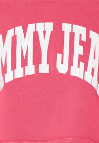 Rosa tröja med ribbad textur, med vit, sliten "TOMMY JEANS" logotyp i en böjd design över framsidan.