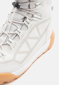 Bota de senderismo ligera en material sintético blanco roto, con un empeine texturizado, sistema de cordones integrado y una suela de goma con una entresuela beige.
