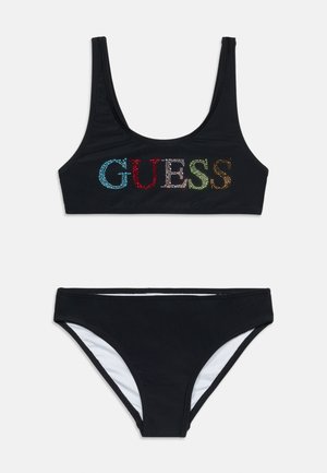 Maillot de bain deux pièces noir avec un haut à encolure arrondie, orné du logo "GUESS" en strass colorés, et un bas uni taille mi-haute avec doublure blanche.