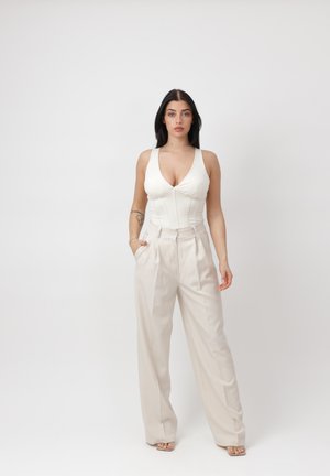 Mujer de cabello largo y oscuro que lleva una blusa blanca sin mangas y pantalones beige de pierna ancha y talle alto, parada con una mano en el bolsillo.