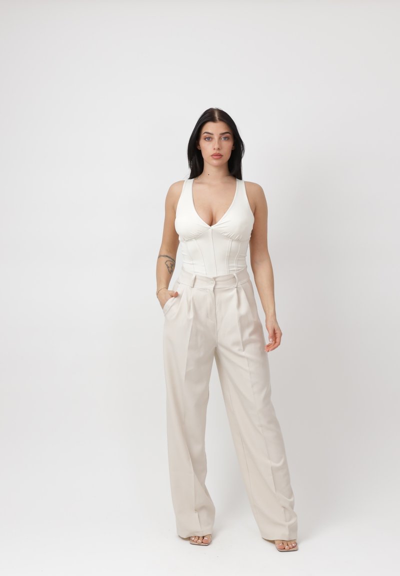 Mujer de cabello largo y oscuro que lleva una blusa blanca sin mangas y pantalones beige de pierna ancha y talle alto, parada con una mano en el bolsillo.