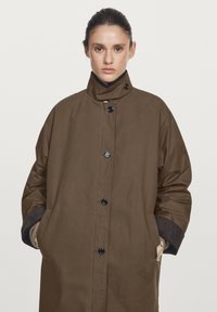 Brown waterproof coat with a high collar, featuring black buttons, side pockets, and contrasting cuffs; smooth texture and oversized design.