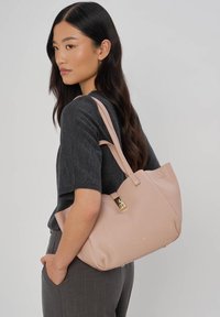 Femme aux longs cheveux bruns portant un haut et un pantalon gris charbon, portant un sac à main en cuir rose clair avec des ferrures dorées à l'épaule.