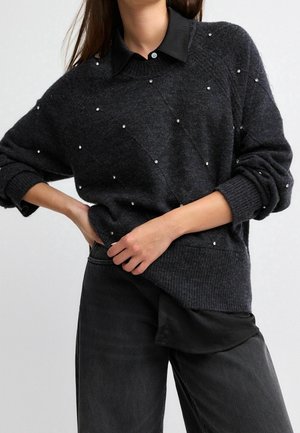 Žena v tmavě šedém svetru s malými bílými korálky přes černou košili s límečkem, v černých kalhotách, ruce opřené o boky.