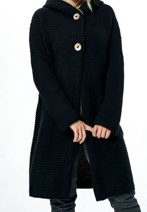 Personne portant un long cardigan noir en tricot avec deux gros boutons en bois, le fermant à l'avant avec les mains.
