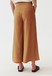 Une personne portant un pantalon marron taille haute et à jambes larges avec des poches arrière et des sandales noires ouvertes à l'avant, se tenant devant un fond blanc uni.