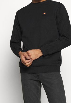 Hombre con una sudadera negra de manga larga con un pequeño logo colorido en el pecho y vaqueros gris oscuro, manos entrelazadas al frente.