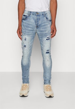 Homme portant un jean skinny bleu clair déchiré avec des éclaboussures de peinture et des baskets beiges, debout devant un fond uni.