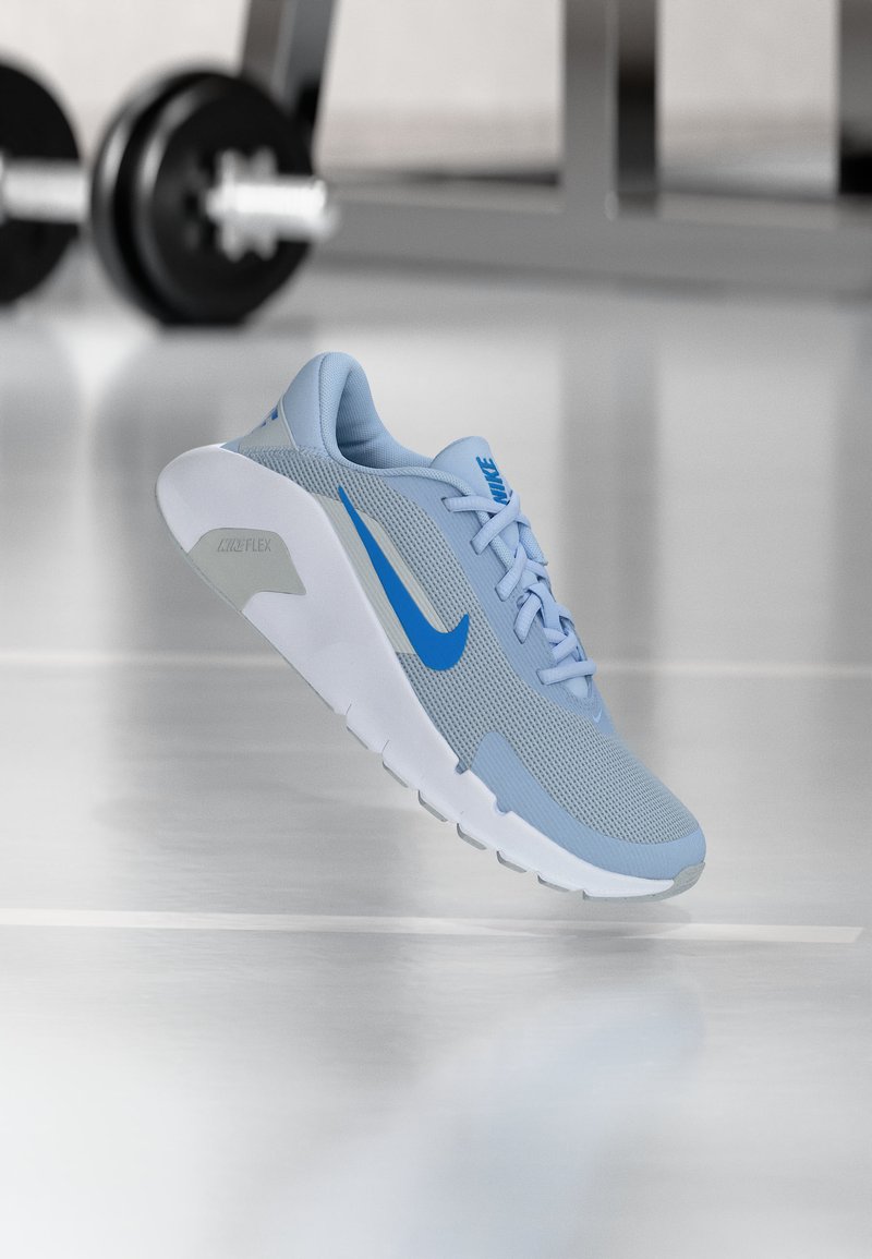 Ανοιχτό μπλε παπούτσι τρεξίματος Nike με λευκή σόλα και μπλε λογότυπο swoosh, τοποθετημένο να αιωρείται πάνω από το πάτωμα του γυμναστηρίου με θαμπά βάρη στο φόντο.