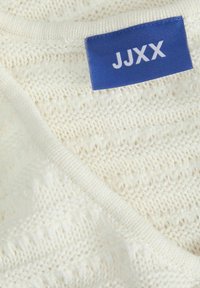 Witte gebreide trui met een textuurpatroon, met een blauwe label waarop de merknaam "JJXX" in het wit is gestikt.