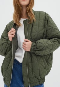 Prošívaná olivově zelená bomber bunda s zipovým uzávěrem, žebrovanými manžetami a kulatým výstřihem, nošená přes bílé tričko a modré džíny.