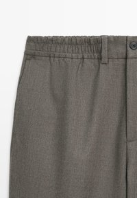 Gros plan d'un pantalon gris sur-mesure avec taille élastique, poches latérales, passants de ceinture et fermeture par bouton.