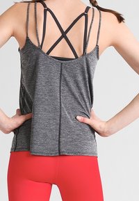 Szary sportowy top na ramiączkach z czarnymi, krzyżującymi się paskami z tyłu, o luźnym kroju i subtelnych przeszyciach, w zestawieniu z czerwonymi legginsami.