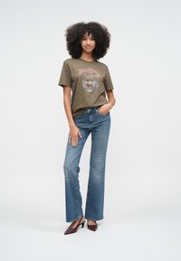 Mujer con cabello rizado que lleva una camiseta gráfica de tigre color oliva, jeans azules con estampado y zapatos de tacón burdeos, de pie sobre un fondo blanco.