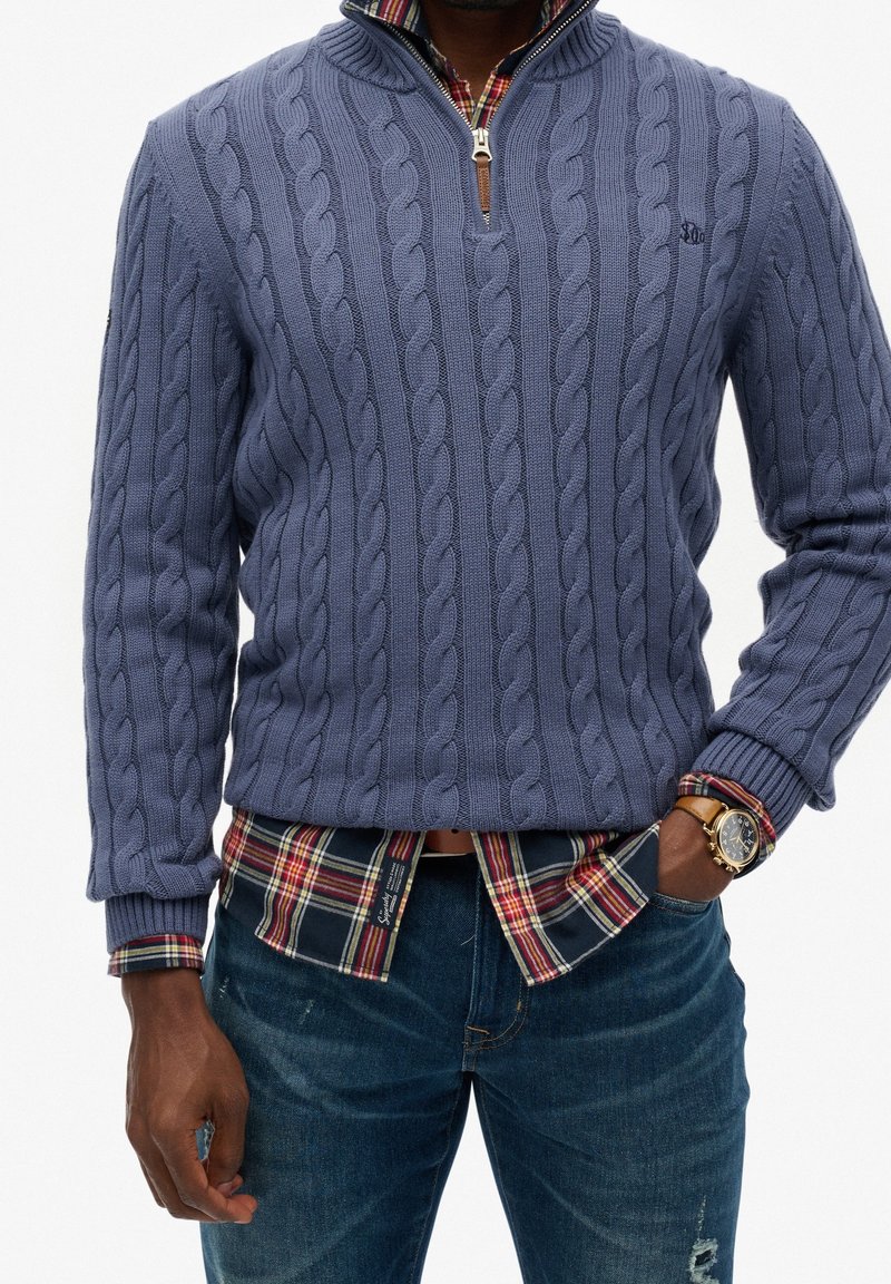 Homme portant un pull bleu à torsades par-dessus une chemise à carreaux rouge avec un jean bleu, main dans la poche, et une montre-bracelet dorée au poignet gauche.