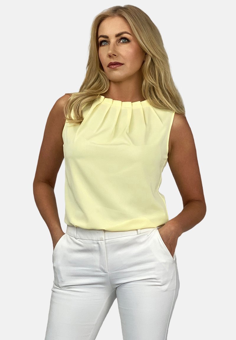 Bezramieniowa żółta bluzka z plisowanym dekoltem, w połączeniu z białymi spodniami. Gładki materiał, luźny krój, czyste linie i minimalistyczny design.