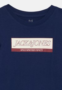 Sötétkék póló, krém és bordó színű téglalap alakú logóval, amely a következő szöveget tartalmazza: "JACK & JONES SINCE NINETEEN NINETY" a mellrészen.