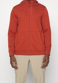 Red zip-up hoodie made of soft fabric. Features a black logo on the chest, drawstring hood, and two front pockets.