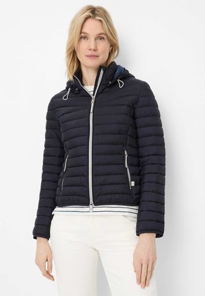 Frau steht und trägt eine schwarze gesteppte Kapuzenjacke mit silbernen Reißverschlüssen, weiße Hose und ein gestreiftes Hemd unter der Jacke.