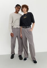 Two individuals stand side by side, wearing loose-fitting gray pleated trousers. One wears a textured cream sweater, the other a black t-shirt with "RAVE" in yellow.