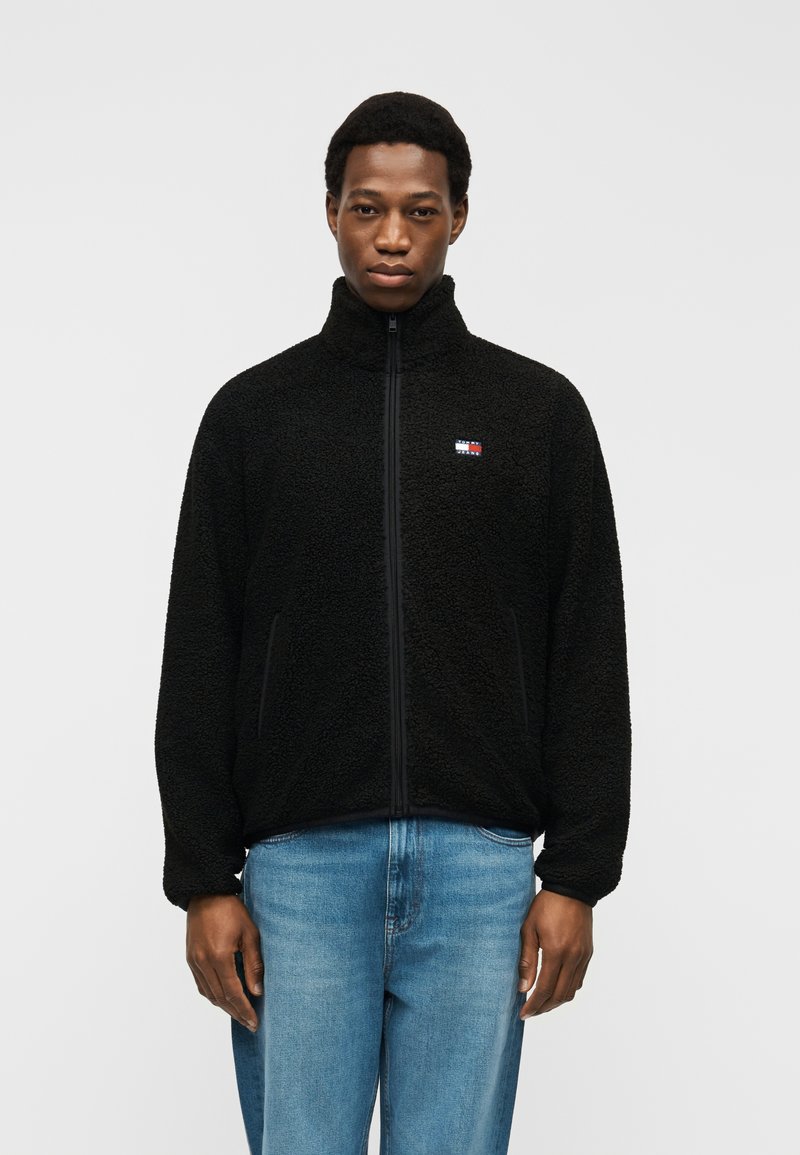 Άνδρας που φοράει μαύρο φλις μπουφάν με φερμουάρ και μπλε τζιν, στέκεται μπροστά σε μονόχρωμο ανοιχτό φόντο, κοιτάζοντας μπροστά με ουδέτερη έκφραση.