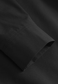 Melnas krekla auduma tuvplāns ar nolocītu aproci, redzamu šuvumu, gludu tekstūru bez rakstiem vai papildus aprīkojuma.