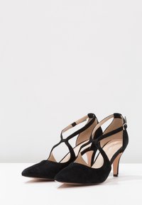 Scarpe nere in suede con tacco alto, punta affusolata e design a T-strap. Presentano una sottile cinghia alla caviglia e un tacco slanciato.