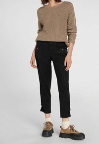 Maglione beige a maglia con scollatura rotonda, abbinato a pantaloni neri a vita alta con tasche frontali e dettagli con bottoni. Scarpe marroni stringate completano il look.