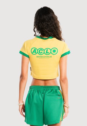 Kvinde med langt krøllet hår iført gul crop top med grøn kant og grønne shorts, stående med ryggen mod på en ensfarvet baggrund.