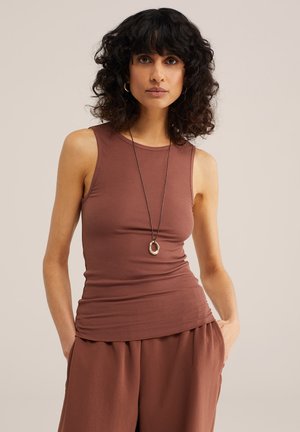 Kobieta o kręconych ciemnych włosach, ubrana w brązowy, bezrękawnik o prążkowanej fakturze i luźne brązowe spodnie, uzupełnione naszyjnikiem z zawieszką i kolczykami-kołami.