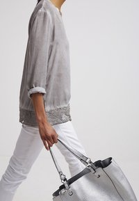 Blouse grise en tissu soyeux avec manches trois-quarts et bordure en dentelle. Associée à un pantalon blanc et un grand sac à main argenté texturé.